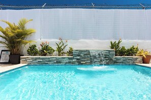 Una piscina con cascada, rodeada de plantas en macetas y una valla blanca.