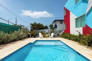 A swimming pool with two white chairs and a small table, surrounded by a green fence and a building with a red and blue facade. - This listing is for two (2) spacious  homes, next to each other (San Juan)
