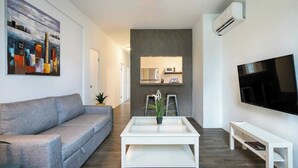 Een moderne woonkamer met een grijze bank, een witte salontafel met een plant, een flatscreen-tv en een keuken met een magnetron en spoelbak op de achtergrond.