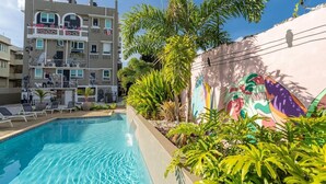 Una piscina con sillones de descanso, un mural y un edificio de varios pisos con balcones.