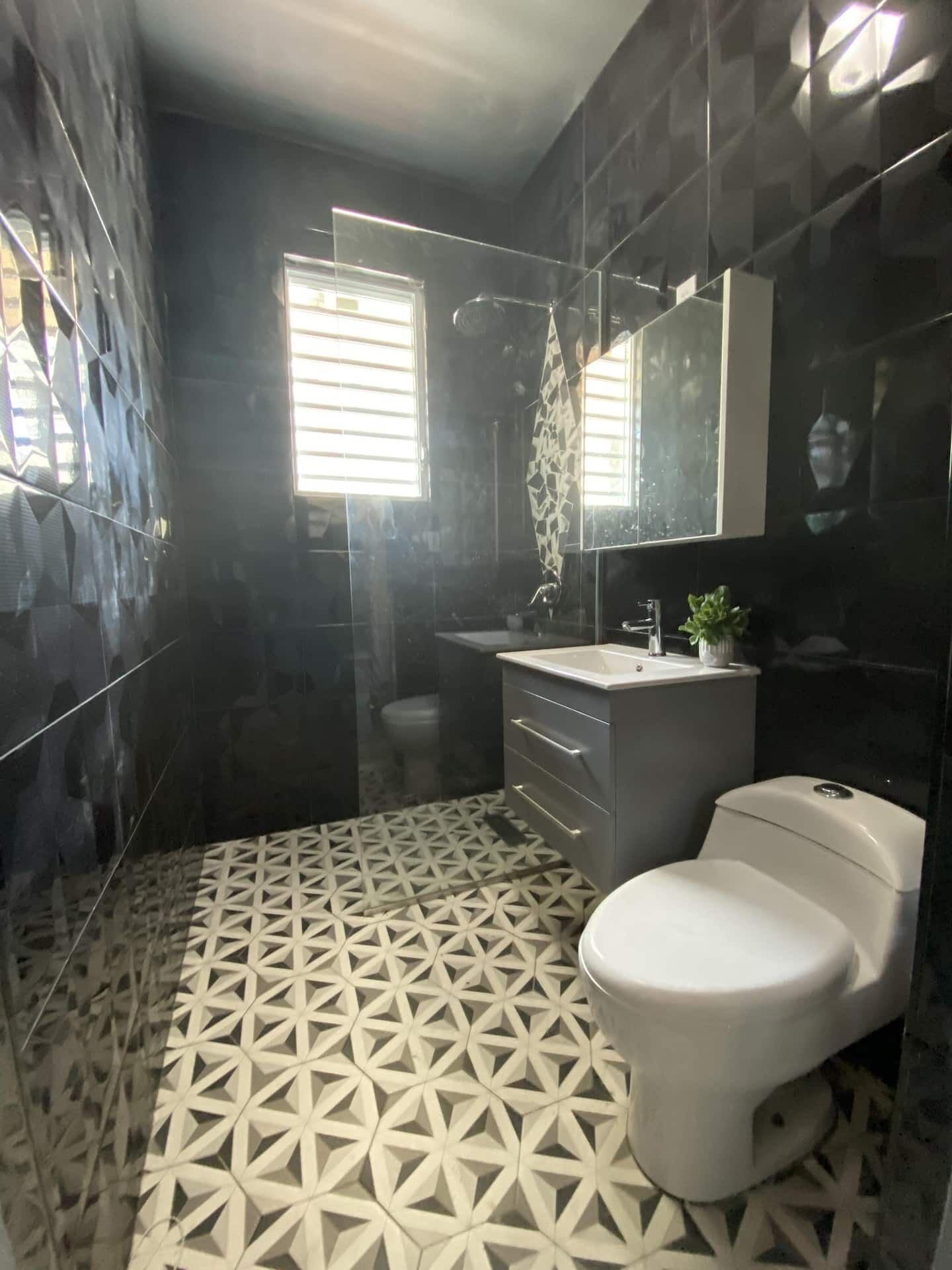 Une salle de bain moderne avec un carrelage au sol à motifs géométriques, des toilettes blanches, un lavabo avec un miroir au-dessus et un espace douche.