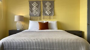 A bed with a textured blanket, two pillows, a bedside lamp, and two wall hangings with geometric patterns.
