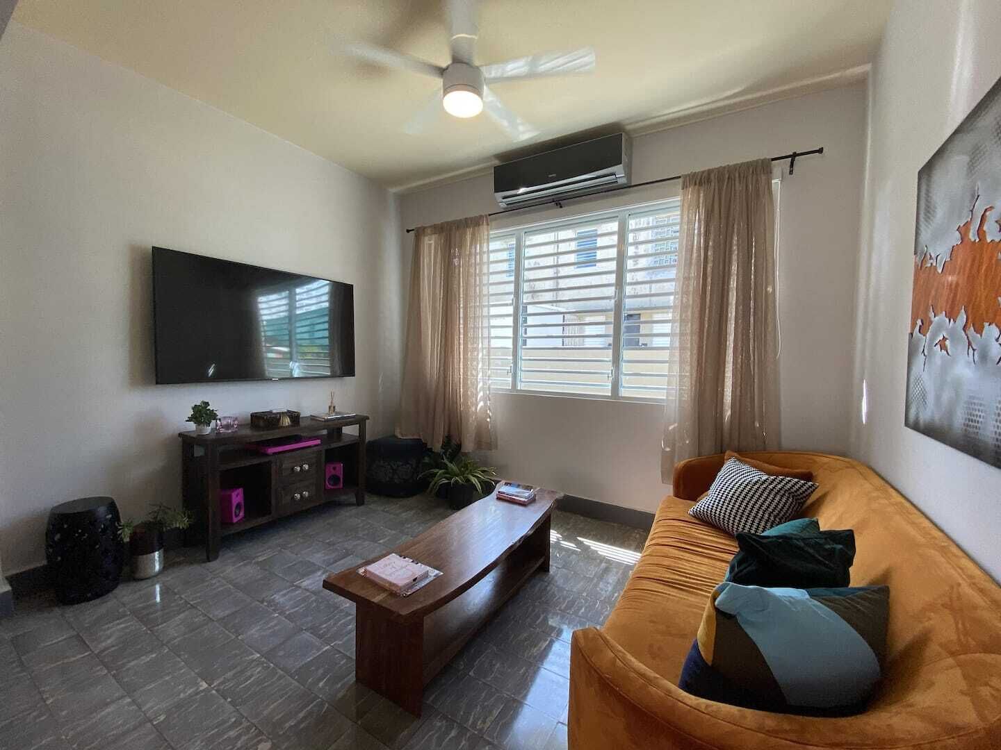 Sala de estar con televisor de pantalla plana, ventilador de techo, ventana con cortinas, sofá con cojines y una mesa de centro.