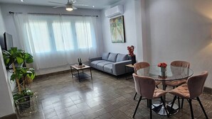 Una sala de estar moderna con un sofá gris, una mesa de centro de vidrio rodeada de sillas rosas, una mesita auxiliar con una planta y una lámpara de pie.