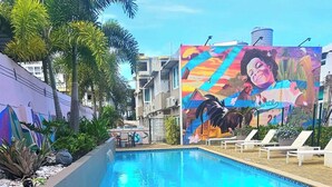 Piscina con un mural, palmeras y sillas de descanso.