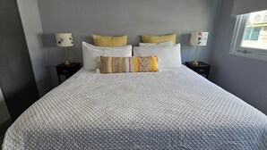 Una cama bien hecha con una colcha blanca acolchada, dos mesitas de noche con lámparas y una ventana con vistas a edificios.
