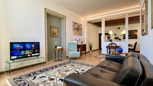 Un salon avec un canapé en cuir, un téléviseur posé sur un meuble en verre, un fauteuil bleu, un tapis à motifs et un coin repas avec une table et des chaises.