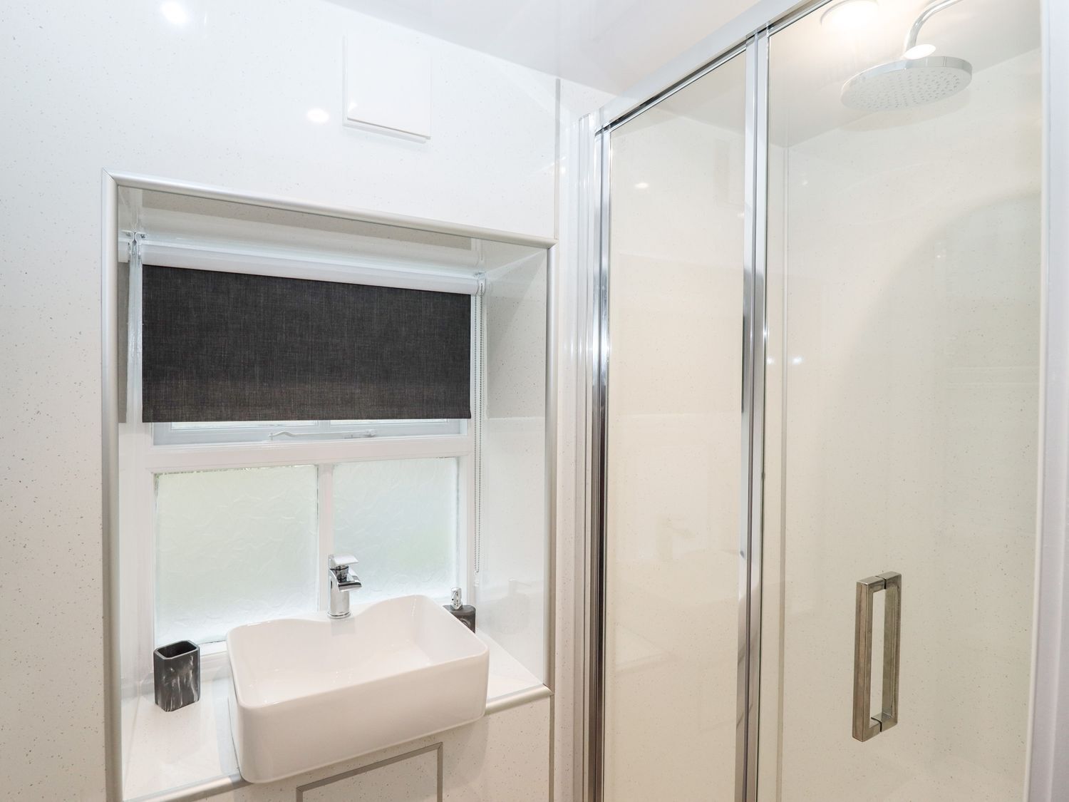 A modern bathroom with a glass shower enclosure, a white sink, and a window with a dark blind.