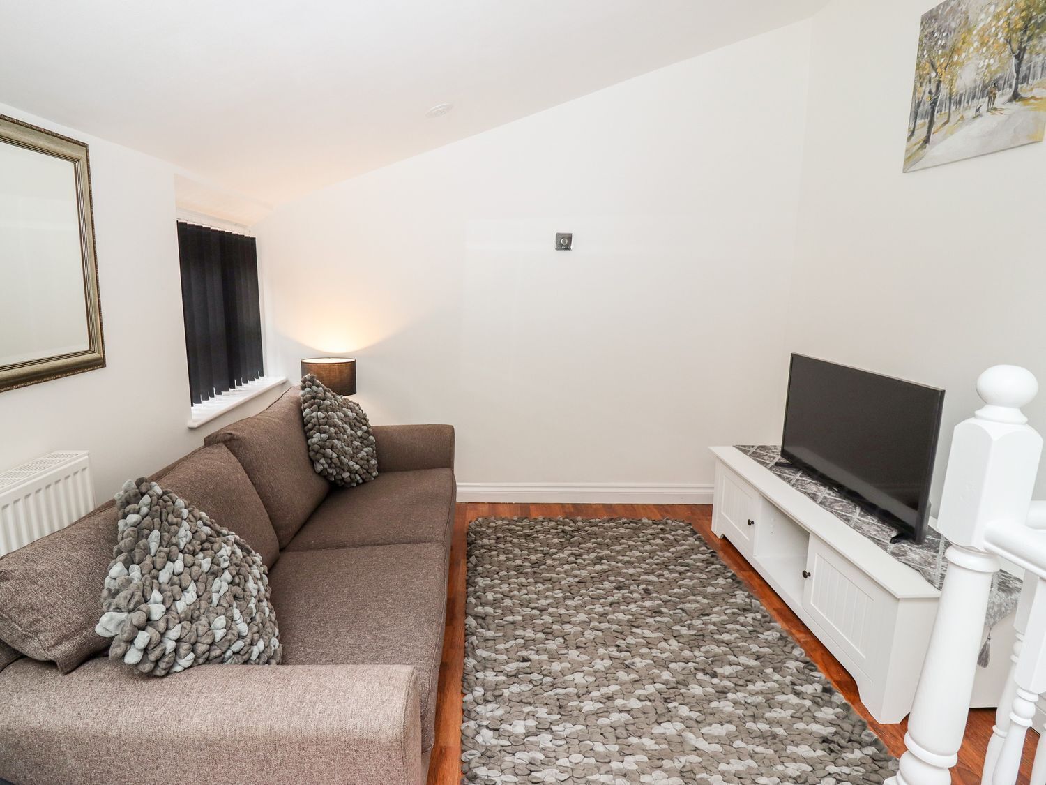 A living room with a brown sofa, a television on a white stand, a pebbled floor rug, and a painting on the wall.