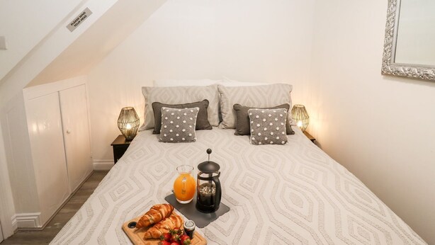 Ein Schlafzimmer mit schrägen Wänden, einem Bett mit gemusterten Betttextilien, zwei Nachttischlampen, einem Nachttisch mit Lampe, einer Ablage mit Croissants und Erdbeeren, einem Glas Orangensaft und einer Kaffeemaschine.