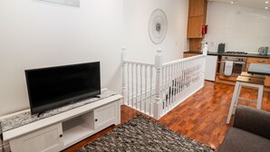 Ein modernes Wohnzimmer mit einem Flachbildfernseher auf einer weißen Unterhaltungseinheit, einem grauen Sofa und einem strukturierten Teppich. Eine Treppe führt zu einer Küchenzeile mit Holzausbau und einem Gaskochfeld.
