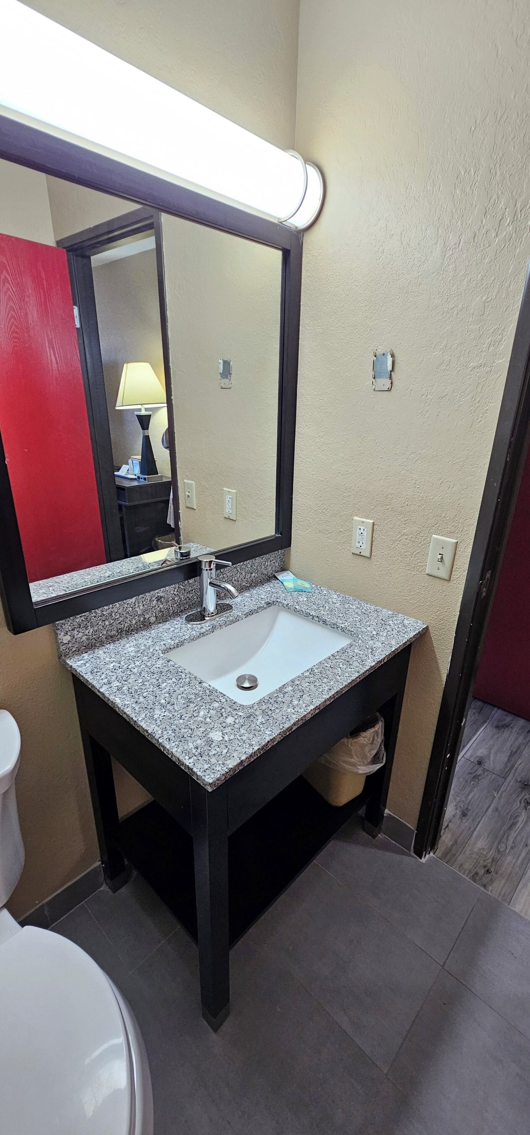 A bathroom with a granite countertop sink, a mirror, a toilet, and a red door.