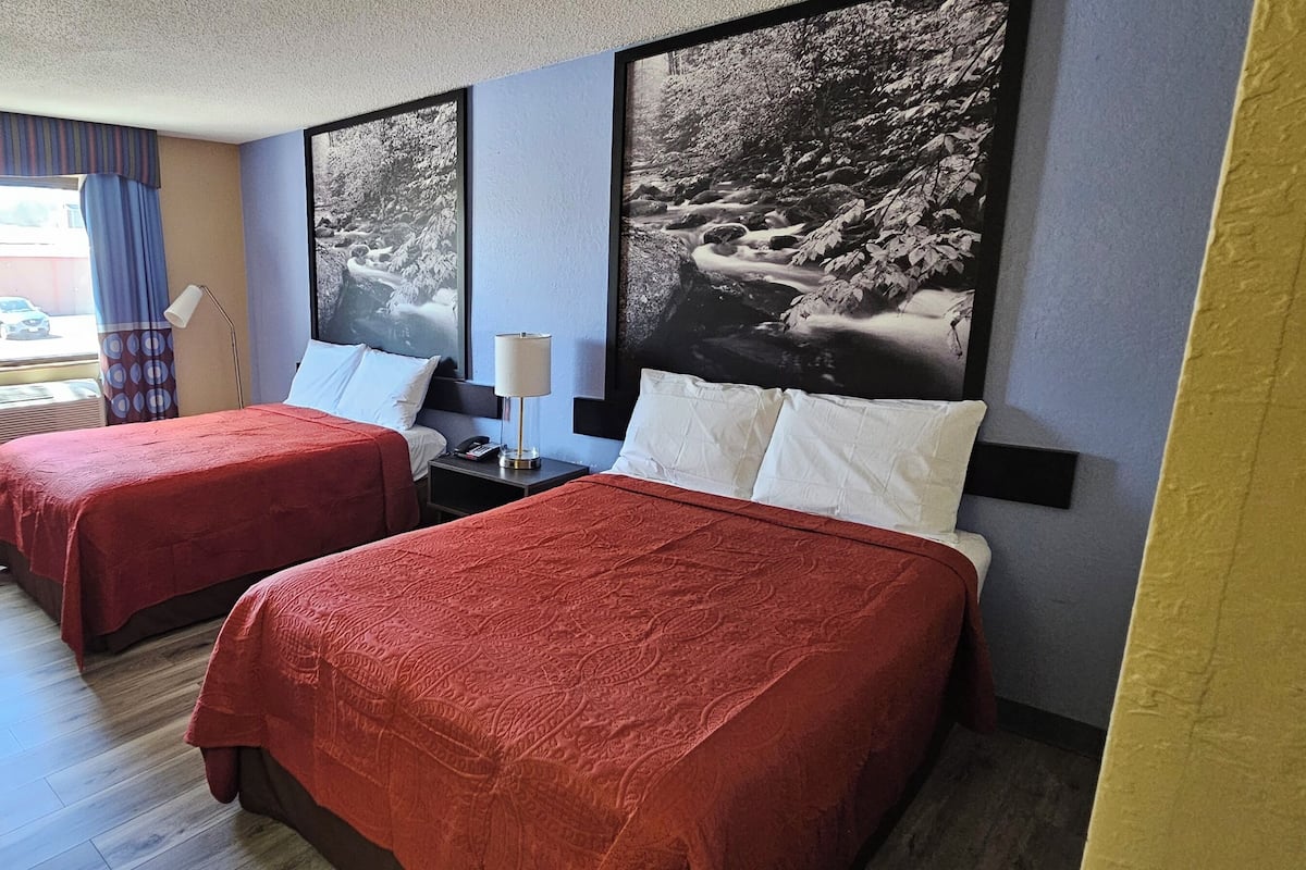 A hotel room with two beds, a desk, a chair, a window with curtains, and two wall-mounted lamps.
