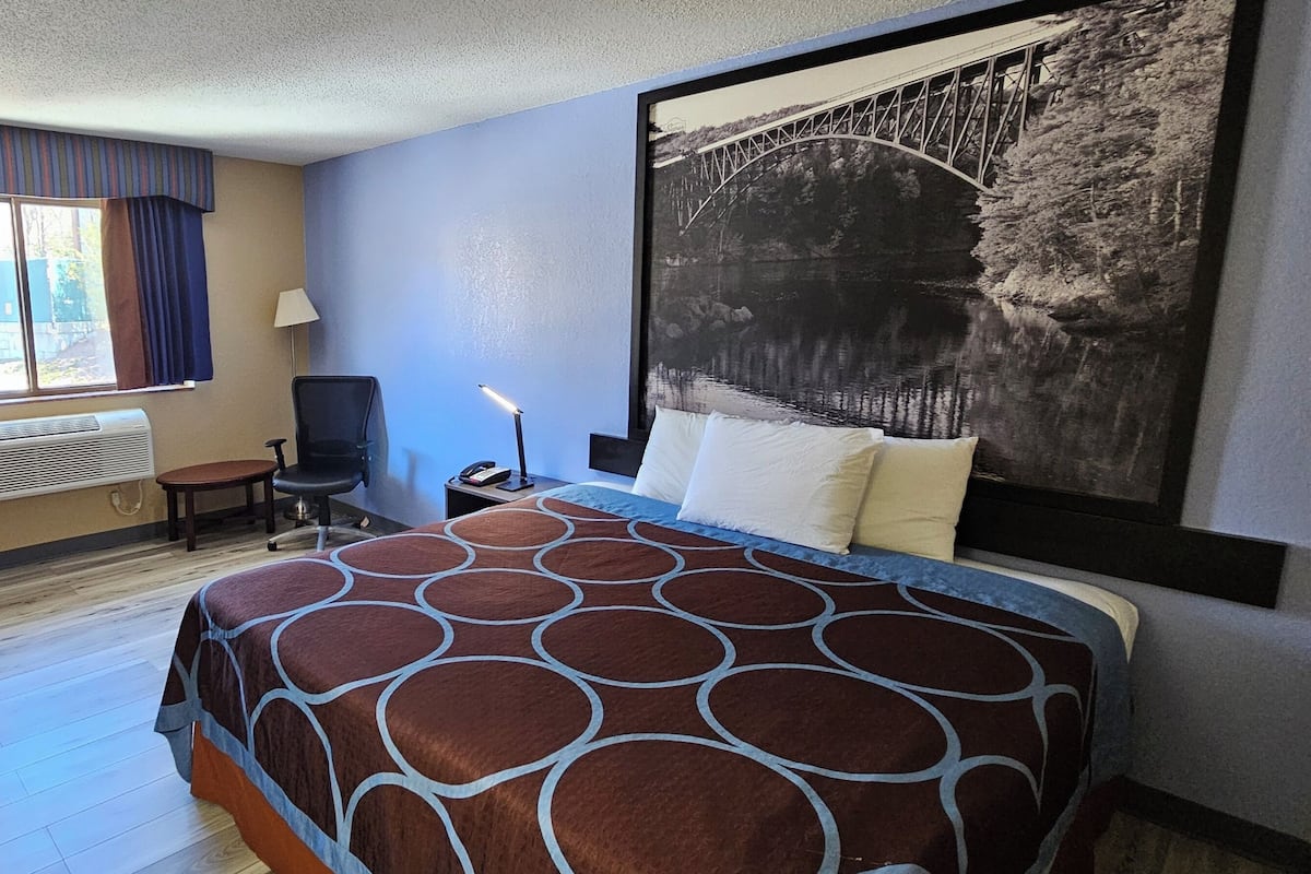 A hotel room with a large bed, a desk with a lamp, a chair, a window with curtains, and a wall-mounted TV.