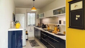 Una cocina compacta con refrigerador, pileta y estufa. Hay una ventana con persianas, una lámpara colgante amarilla y un certificado en el pared.