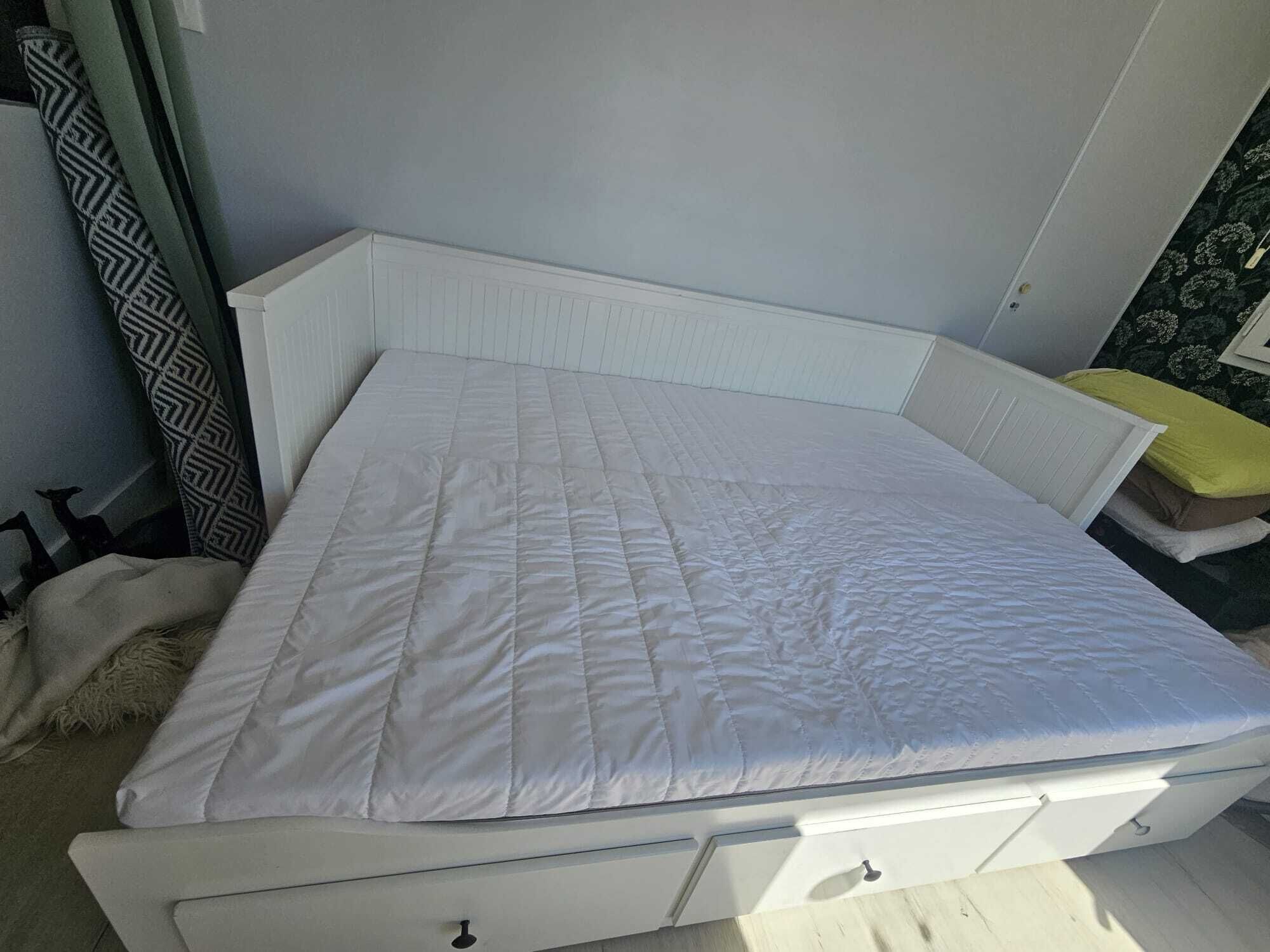 A white bed with a quilted mattress in a room with light-colored walls and a patterned curtain.