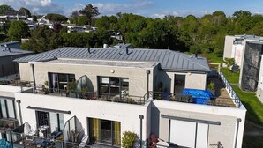 A modern two-story building with a flat roof, large windows, and a balcony with outdoor seating.