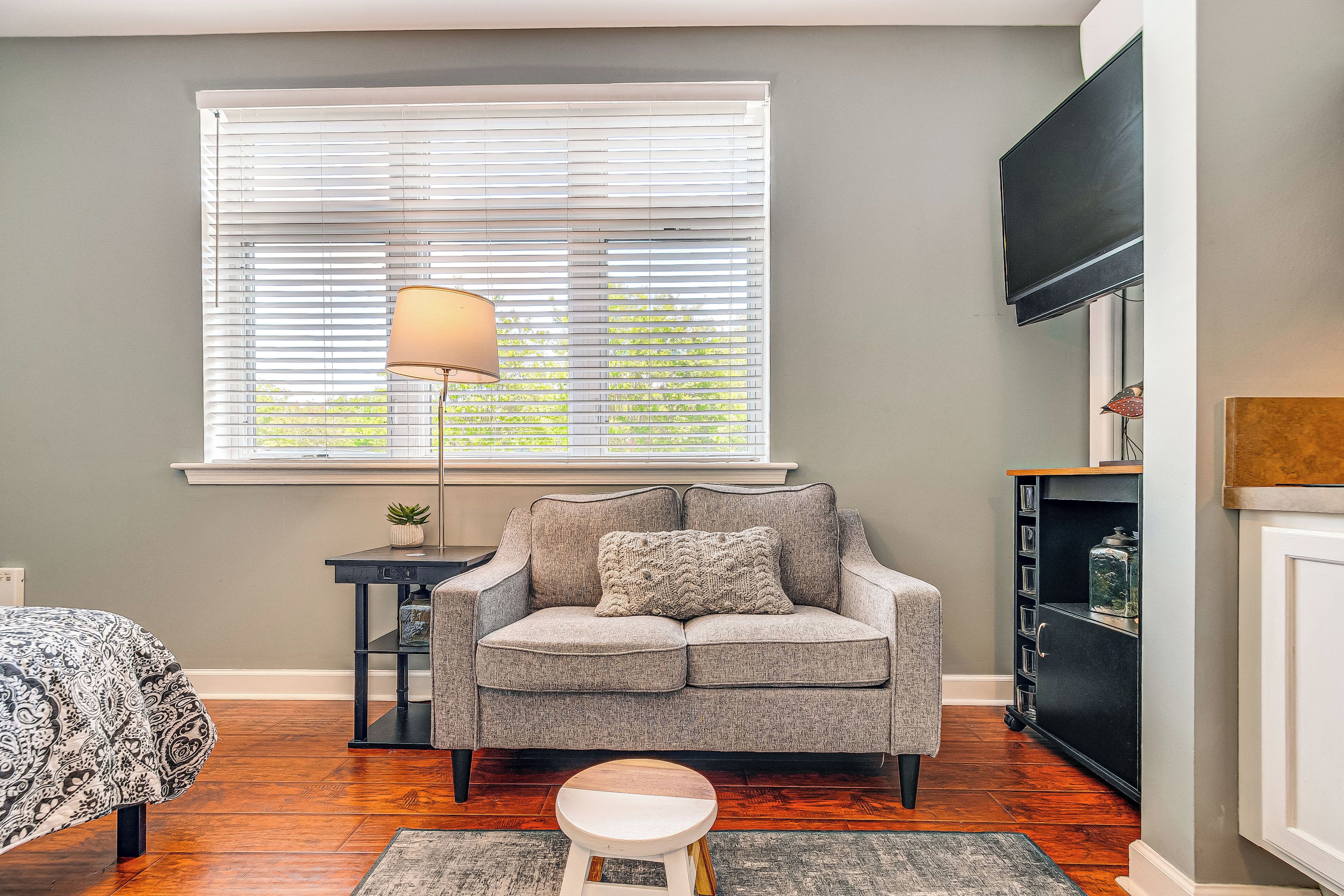A room with a grey sofa, a small round table with a lamp, a bookshelf, and a TV mounted on the wall.
