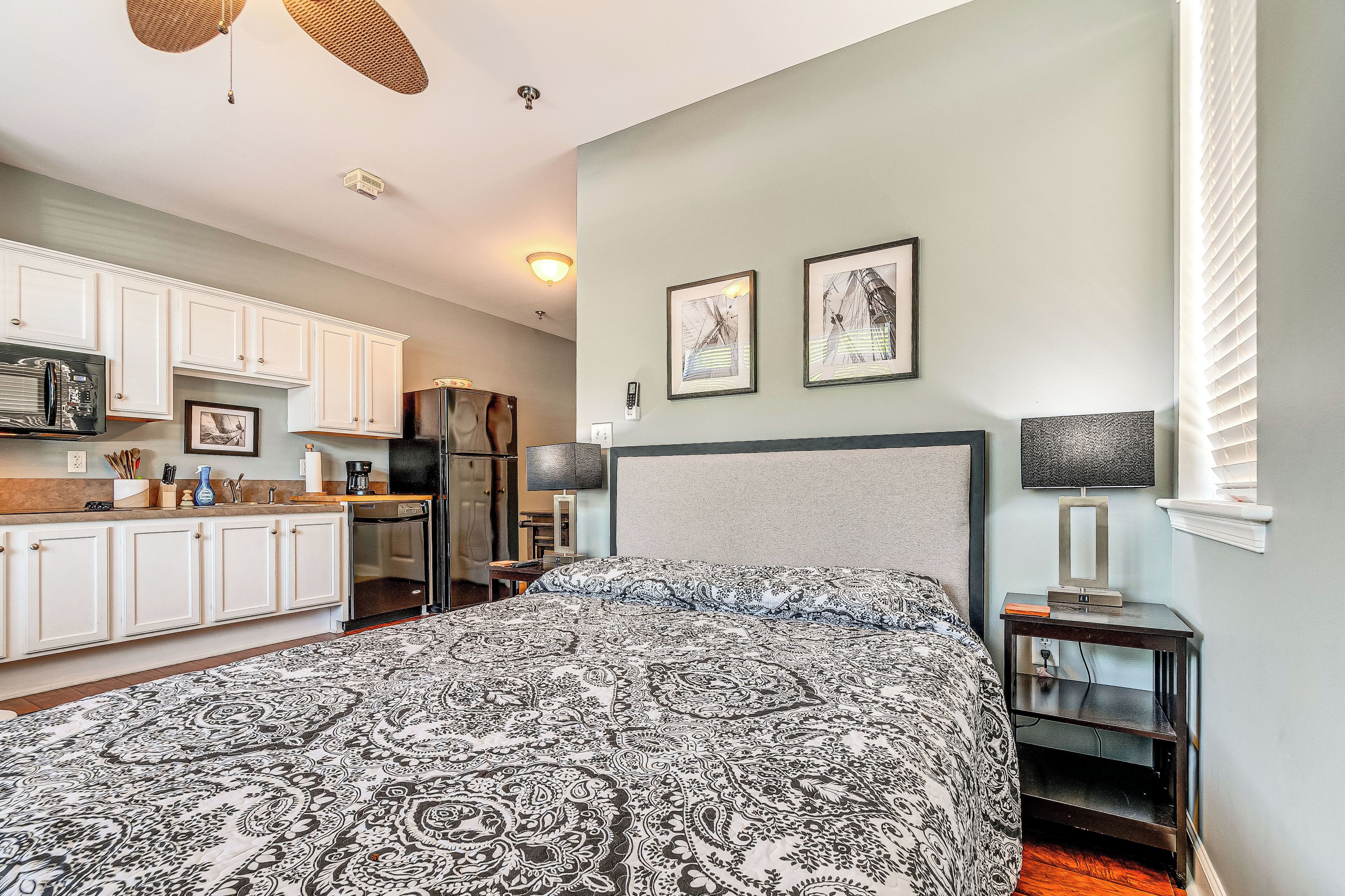 A bedroom with a patterned bedspread, a bedside table with a lamp, and a view of the kitchen area.