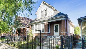 A two-story house with a brick facade and white trim, a front yard with a black metal fence, and a clear blue sky. - Logan Square Urban Oasis: 2 Mi to Local Hotspots! (Chicago)
