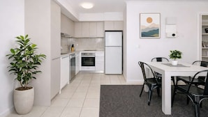 A modern kitchen with white cabinets, a refrigerator, and a dining area with a table and chairs. - Stylish Townsville CBD Apartment with Pool, Parking & Balcony (Townsville City)