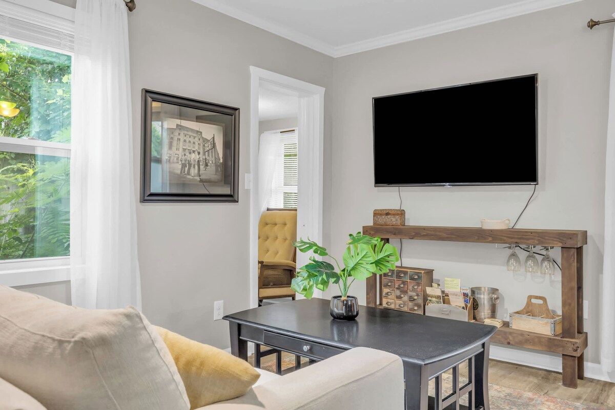 Een woonkamer met een bank, een salontafel, een aan de muur bevestigde televisie en een houten plank met diverse spullen.
