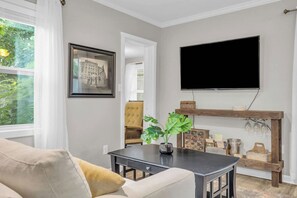 A living room with a sofa, a coffee table, a television mounted on the wall, and a wooden shelf with various items.