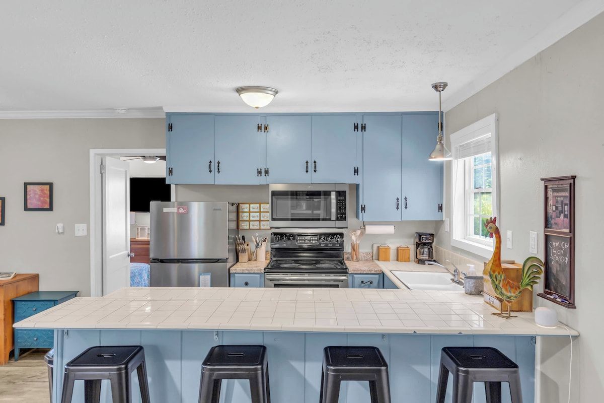 A kitchen with blue cabinets, a white countertop, a stainless steel refrigerator, a microwave, an oven, and a sink. There are four stools at the island.