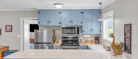 A kitchen with blue cabinets, a white countertop, a stainless steel refrigerator, a microwave, an oven, and a sink. There are four stools at the island.