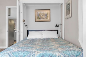 A bedroom with a patterned bedspread, a framed picture on the wall, and a door leading to another room.
