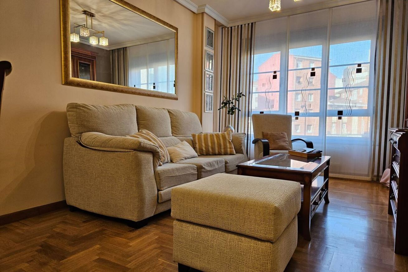 A living room with a sofa, ottoman, and armchair, a large mirror, and a view of buildings through a window.
