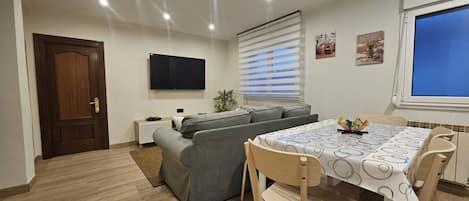 Una sala de estar moderna con un sofá, una mesa de comedor con sillas, un televisor montado en la pared y una ventana con persianas.