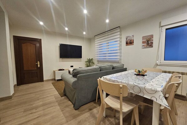 Een moderne woonkamer met een bank, een eettafel met stoelen, een aan de muur bevestigde televisie en een raam met zonwering.