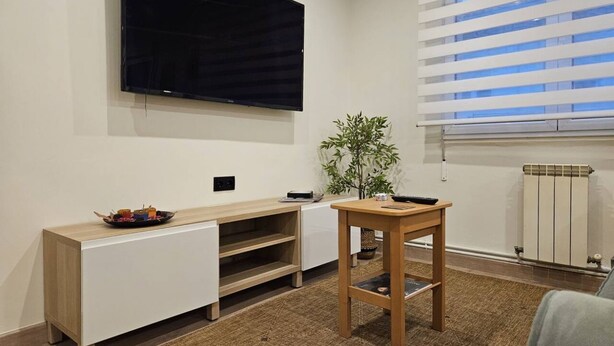 A modern living room with a flat-screen TV mounted on the wall, a wooden side table, a white entertainment unit, and a window with blinds. - 1A111 Apartamento moderno de 3 dormitorios. (Gijón)