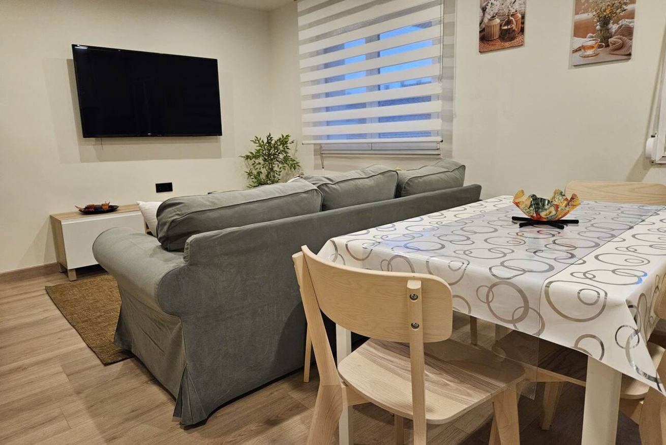 A modern living room with a sofa, a dining table with a patterned tablecloth, a TV mounted on the wall, and a window with blinds.