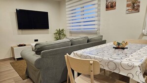 A modern living room with a sofa, a dining table with a patterned tablecloth, a TV mounted on the wall, and a window with blinds. - 1A111 Apartamento moderno de 3 dormitorios. (Gijón)