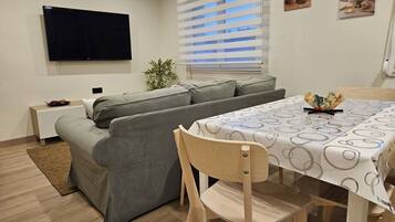 A modern living room with a sofa, a dining table with a patterned tablecloth, a TV mounted on the wall, and a window with blinds.