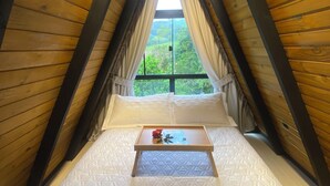 A cozy attic bedroom with a wooden sloped ceiling, a single bed with a quilted mattress, a bedside table with a rose, and a view of greenery outside. - Miraflores Recanto (Imarui)