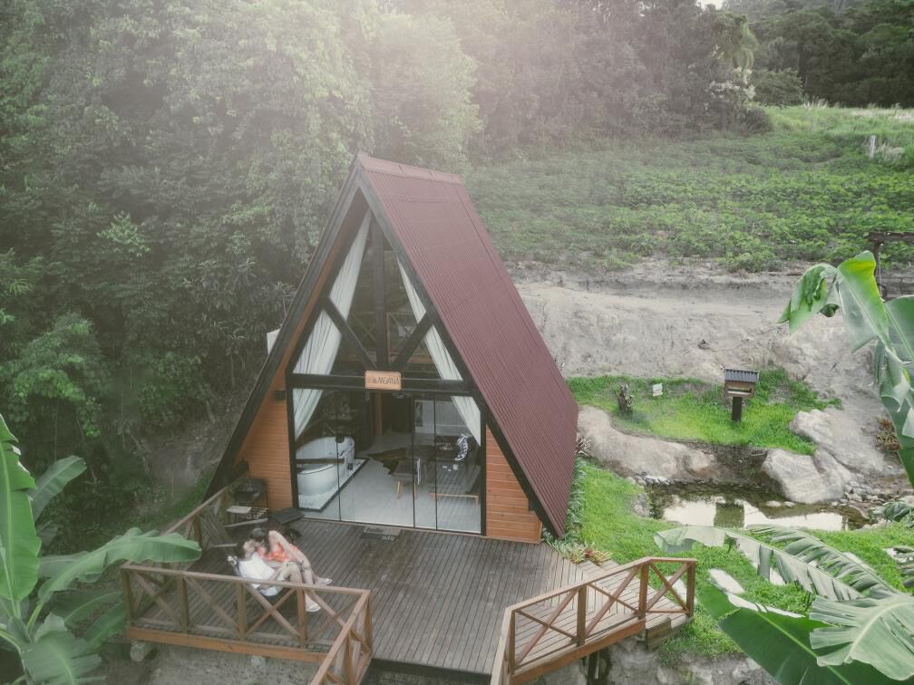 A triangular wooden structure with a glass front, surrounded by lush greenery and a small pond.