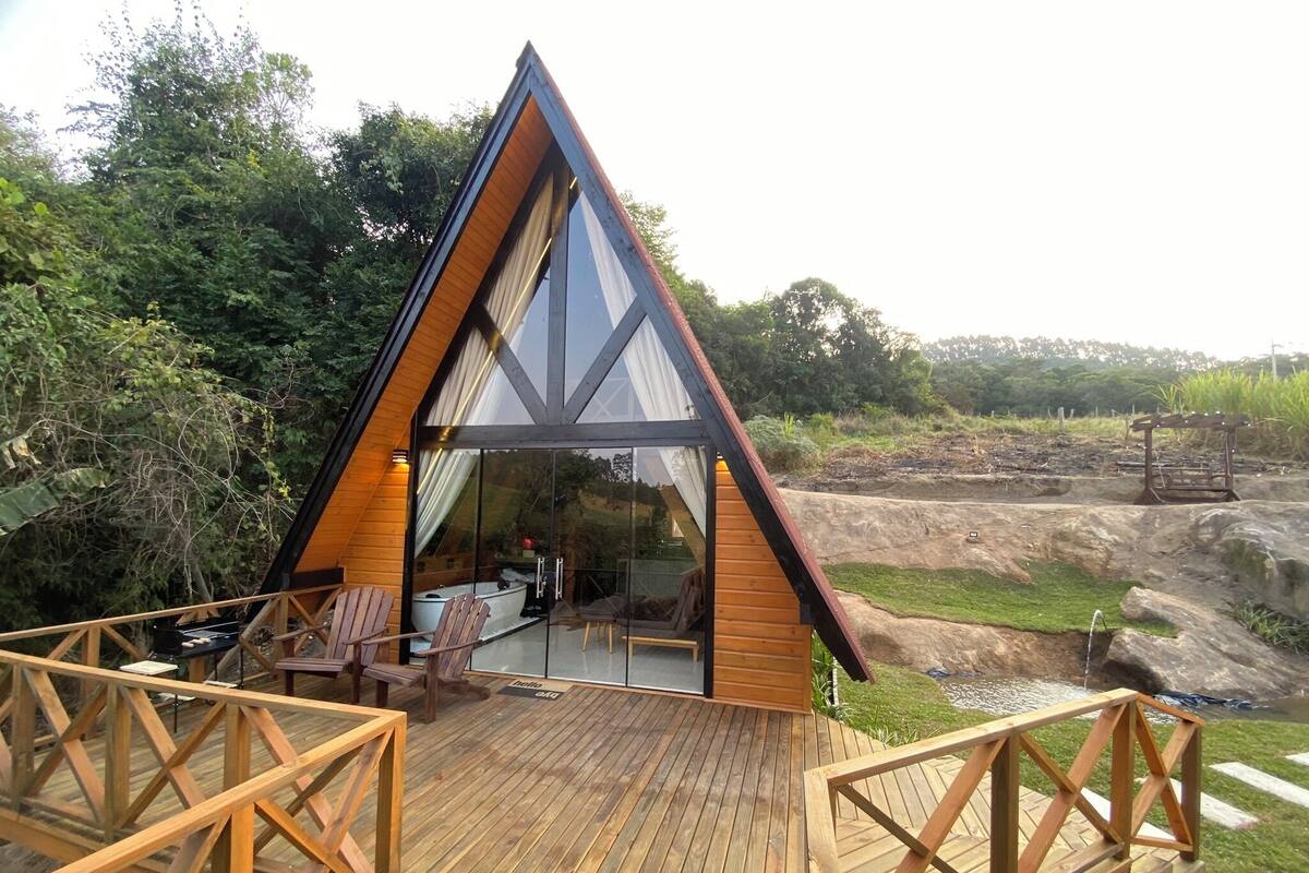 A modern A-frame wooden cabin with a glass balcony, surrounded by lush greenery.
