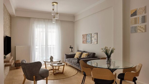 A modern living room with a sofa, coffee table, and chairs. There is a television mounted on the wall and a window with curtains. - Athens Glow 2bd Apt in Ilisia (Athina)