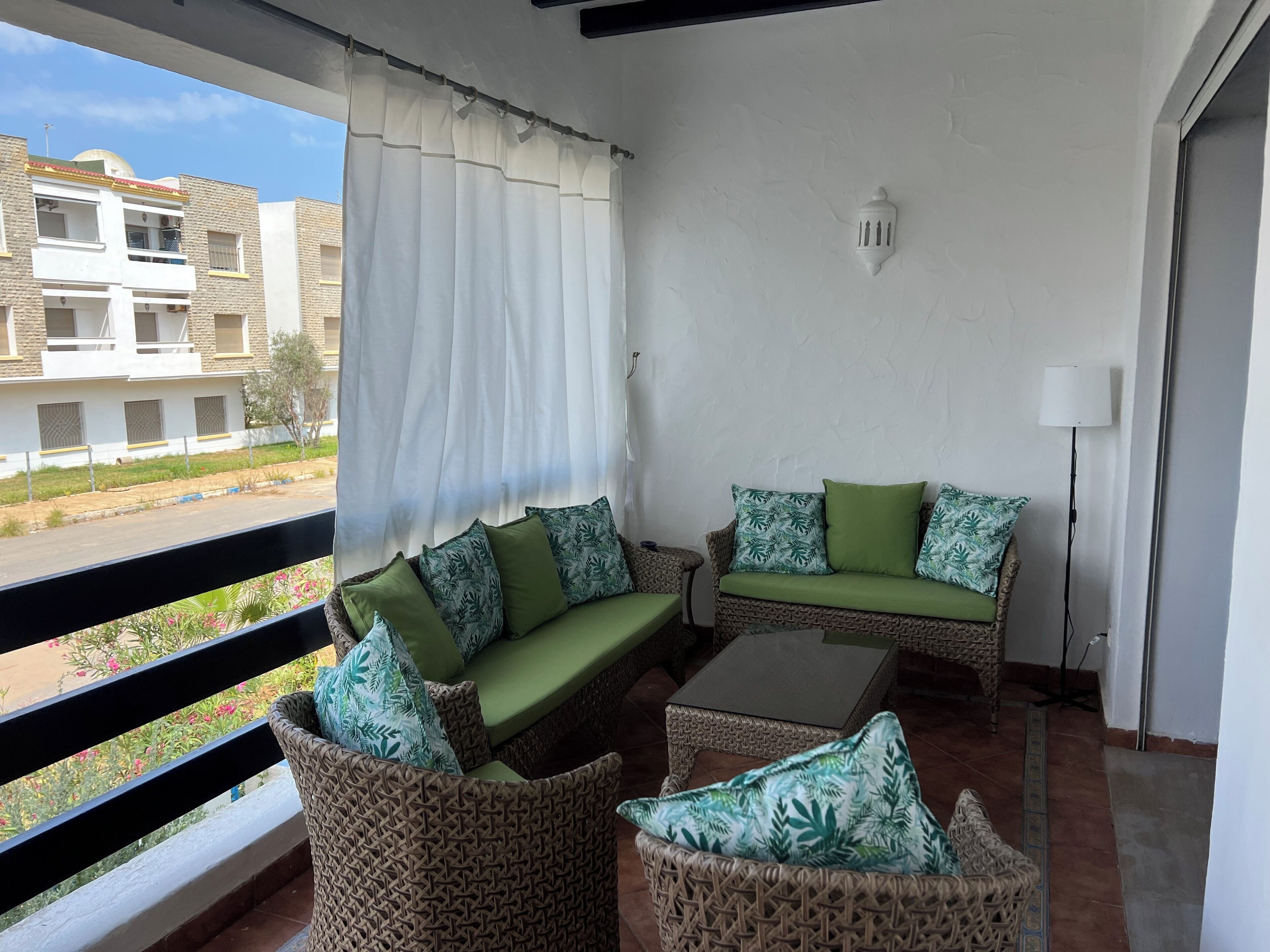 A balcony with wicker furniture, a white curtain, and a view of buildings in the background.