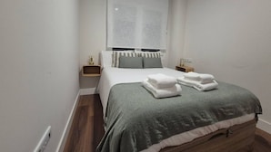 Un dormitorio con cama, ventana con persianas, mesitas de noche y suelo de madera.