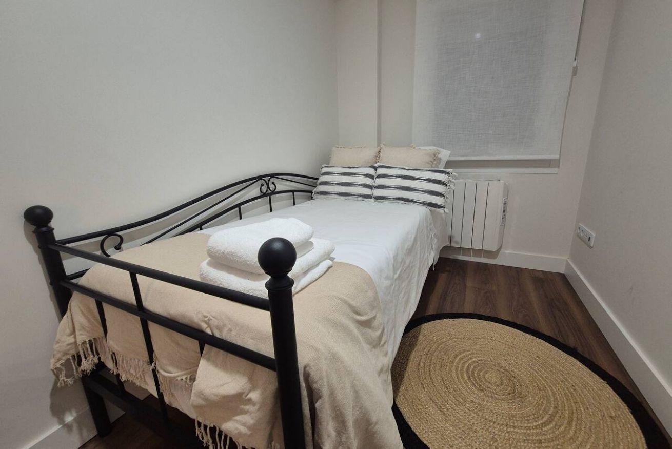 A single bed with a black metal frame, white bedding, and a beige throw blanket. There is a window with a white blind, a radiator, and a woven rug on the floor.