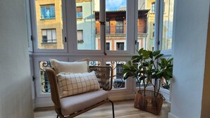 A corner room with a wicker sofa, a potted plant, and a view of neighboring buildings through large windows.