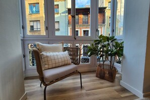 Habitación de esquina con sofá de mimbre, planta en maceta y vista a edificios cercanos a través de amplias ventanas.