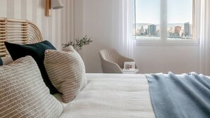A hotel room with a bed, a chair, a window with a city view, and a wall-mounted lamp.