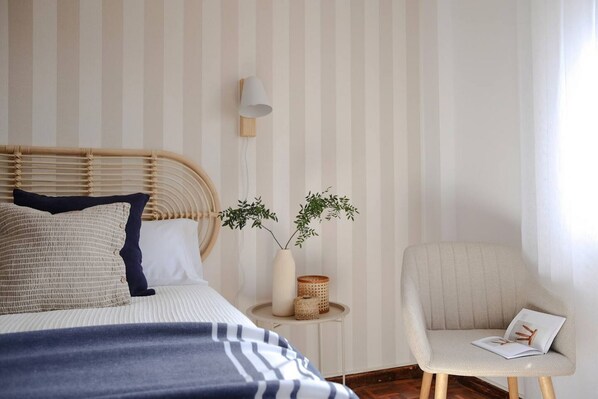 A bedroom with a striped wallpaper, a bed with a headboard, a bedside table with a lamp, a chair, and a plant.