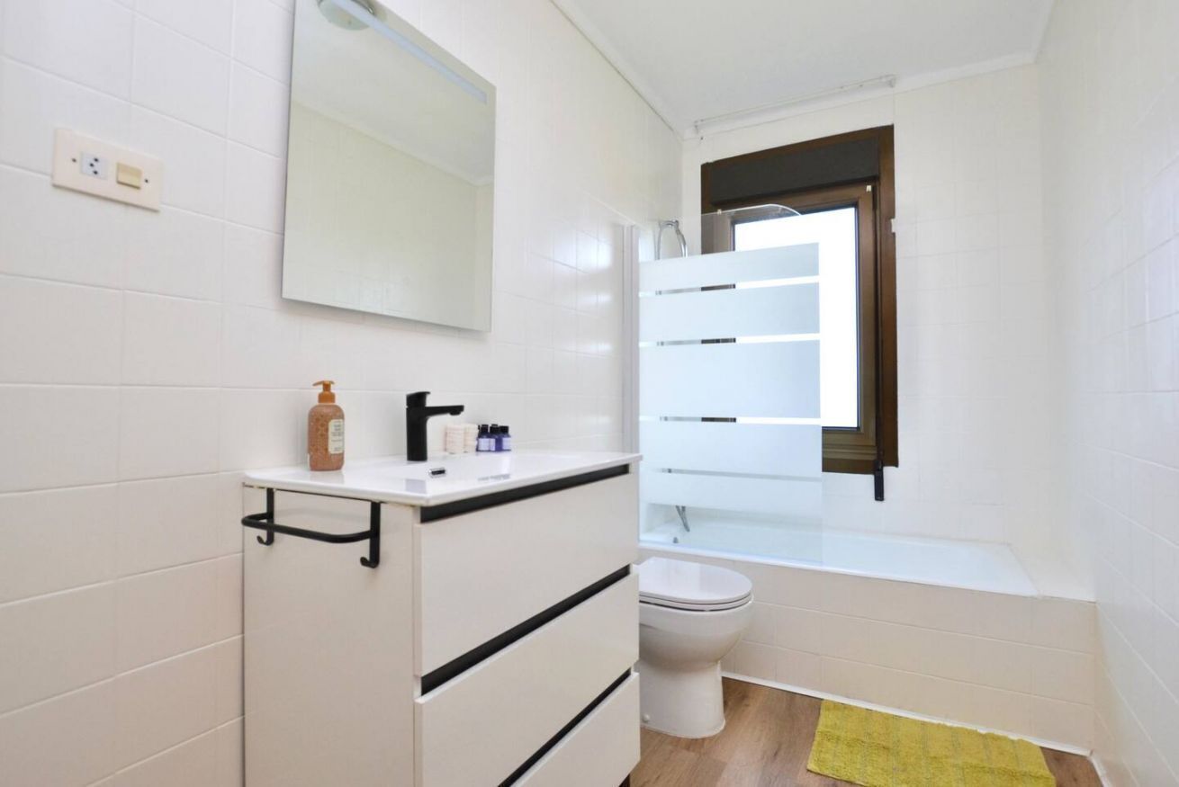 Een badkamer met een wit wastafelmeubel, spiegel en toilet, een geel badmatje en een raam met zonwering.