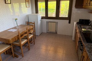 Una cocina con una mesa de comedor, sillas, un refrigerador, una ventana con vista a follaje y un radiador.